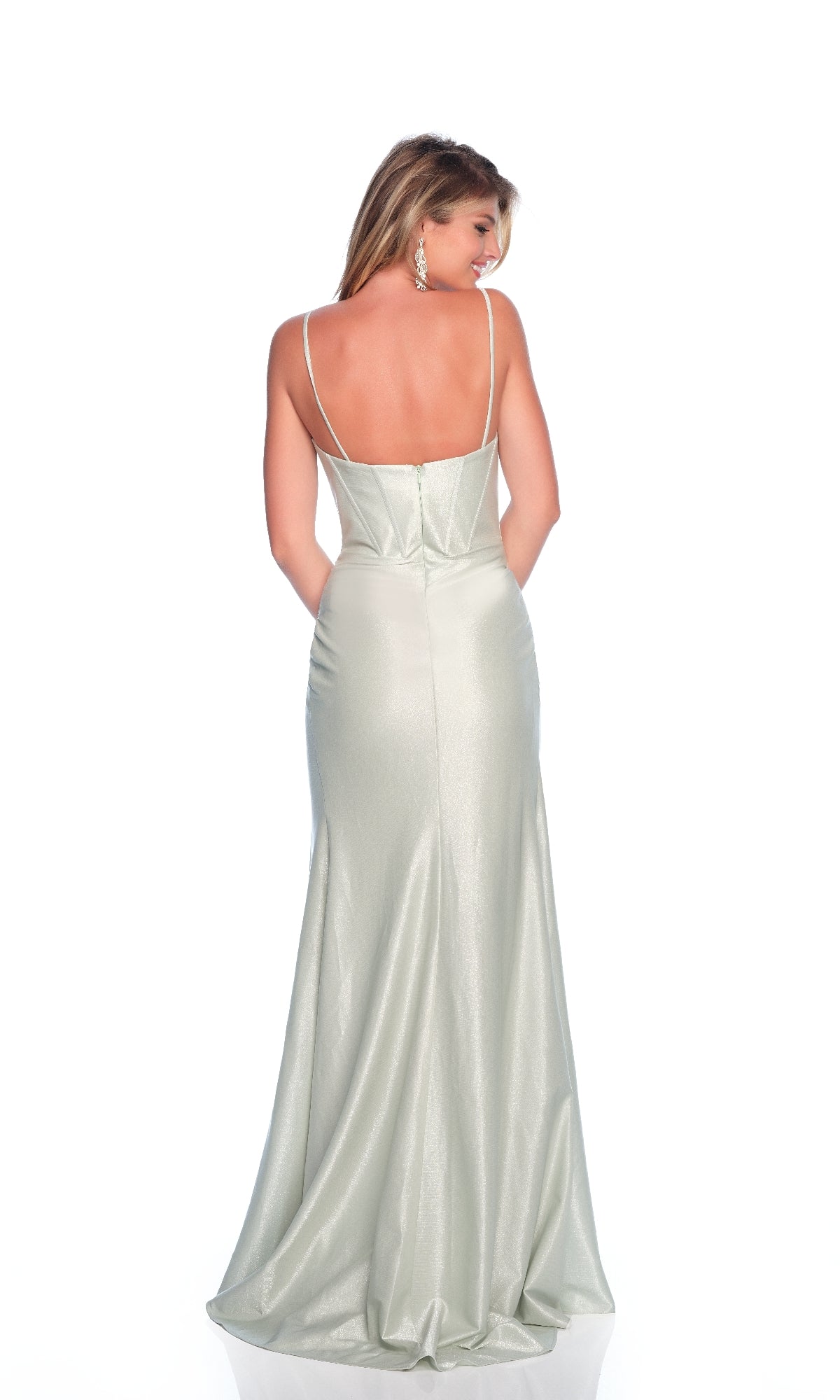 A woman stands facing away against a white background, wearing the Dave and Johnny 12190 long prom dress—a fitted, light silver, floor-length gown with thin straps and a low back.